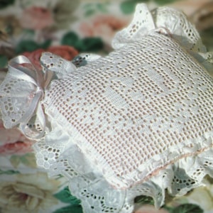 Könnte beinhalten: Ein weißes, quadratisches Häkelkissen mit einem filigranen Lochmuster. Das Kissen hat eine gerüschte Spitzenborte und eine dekorative Schleife aus weißem Band. Ein weiches, romantisches Dekorationselement.