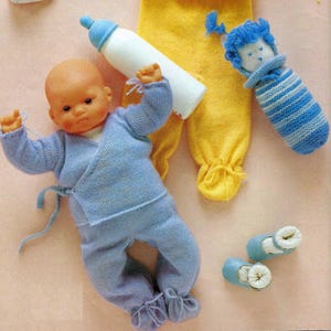 May include: A knitted baby doll outfit in light blue with a matching hat and booties. The outfit has a wrap-style top and a pair of pants. The doll is lying on a pink surface with a bottle, a knitted toy, and a pair of shoes.