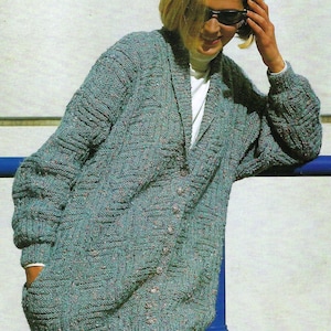 May include: A teal-coloured, long-sleeved cardigan with a textured knit pattern. The cardigan has a V-neck, button closure, and pockets. The garment is approximately knee-length and appears to be made of a soft, warm material. The cardigan is worn over a white top.