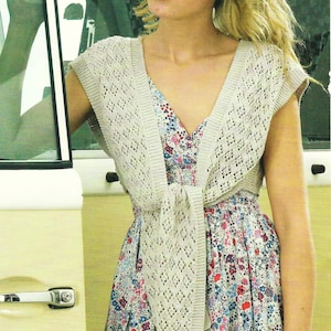 May include: A woman wearing a white crocheted sleeveless cardigan tied at the waist over a floral print dress. The cardigan has a delicate openwork pattern.