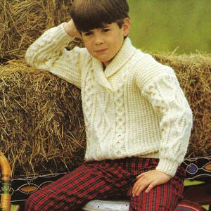 May include: A cream-colored cable-knit sweater with a shawl collar and red plaid pants. The sweater features textured patterns on the front and sleeves. The pants have a red and black plaid pattern. The outfit is suitable for children's clothing.