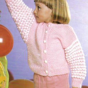 May include: A pink knitted cardigan with white buttons and bobble stitch detailing on the sleeves. The cardigan is worn by a child with blonde hair.