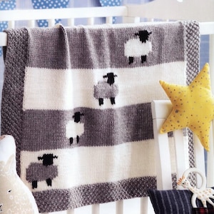 Pode incluir: Um cobertor de bebé de malha com um padrão de riscas em cinzento e branco. O cobertor apresenta um desenho repetido de ovelhas com faces pretas. O cobertor está drapeado sobre um berço branco.