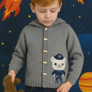 May include: A grey hooded cardigan with wooden buttons and a bear design. The bear wears a blue uniform. The child wears blue jeans and holds a brown teddy bear. The background features a space theme with a rocket and planets.