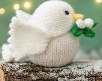 May include: A knitted white bird figurine with a yellow beak and black eyes, holding a sprig of green leaves and white berries. The bird is perched on a snow-covered log, with a blurred background of soft lights.