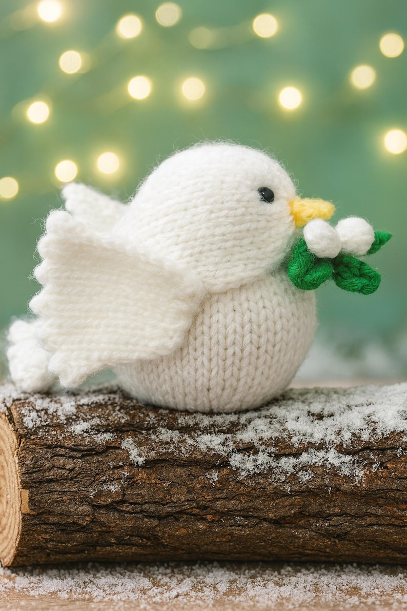 May include: A knitted white bird figurine with a yellow beak and black eyes, holding a sprig of green leaves and white berries. The bird is perched on a snow-covered log, with a blurred background of soft lights.