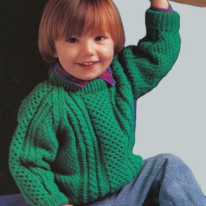 Könnte beinhalten: Ein Kind trägt einen grünen Strickpullover mit strukturiertem Muster und einem karierten Kragen. Der Pullover hat einen Rundhalsausschnitt und lange Ärmel. Das Kind trägt auch eine blaue Cordhose.