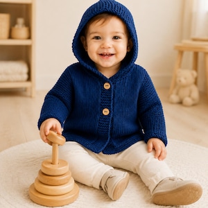 May include: A deep blue knitted hooded cardigan featuring two light-colored wooden buttons on the front. The garment has long sleeves and a textured knit pattern, providing a warm layer for a child, paired with light-colored pants and shoes.