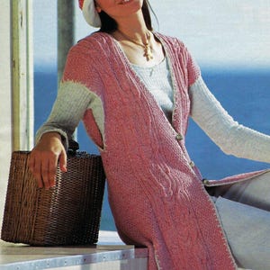 May include: A woman wearing a pink cable knit vest with a white trim, a white hat with a red ribbon, and a white shirt. She is holding a brown wicker basket.