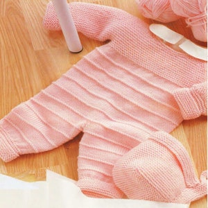 May include: A light pink knitted baby outfit, including a long-sleeved onesie with ribbed detailing and a matching cap. The outfit is made of soft yarn and is displayed on a wooden surface, with balls of yarn in the background.