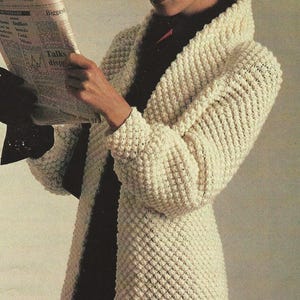May include: A white crocheted cardigan with a wide collar. The cardigan is made with a textured stitch that creates a raised, bumpy effect. The model is wearing a black shirt underneath the cardigan.