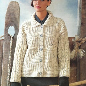 Puede incluir: Un cárdigan blanco de punto con un patrón de punto de cable y cierre de botones. El cárdigan tiene cuello y se lleva con un gorro gris y guantes negros.