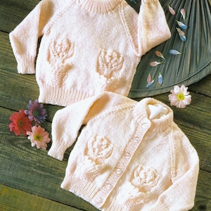 May include: Two pale pink knitted baby sweaters. One is a crew neck, the other a button-up cardigan. Both feature floral designs on the front. The sweaters are on a green wooden surface with flower petals.