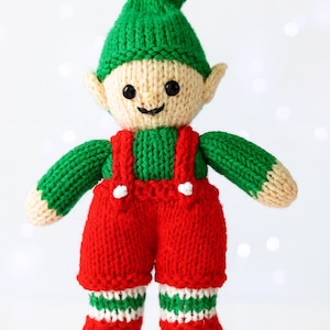 Könnte beinhalten: Eine handgestrickte Elfenpuppe mit grünem Hut und Armen, roter Latzhose und gestreiften Socken. Die Puppe hat ein beiges Gesicht mit schwarzen Knopfaugen und einem kleinen Lächeln. Eine rote Glocke schmückt den Hut. Eine festliche Weihnachtsdekoration.