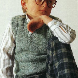 Puede incluir: Una persona con un chaleco de punto gris sobre una camisa a rayas, combinado con pantalones a cuadros. La persona también lleva gafas con montura marrón. El atuendo tiene una estética vintage.
