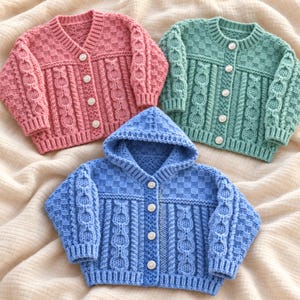 May include: Three knitted cardigans in pink, mint green, and blue. The pink and green cardigans have round necklines, while the blue cardigan features a hood. All garments display a textured cable knit pattern, ribbed cuffs, and white buttons.