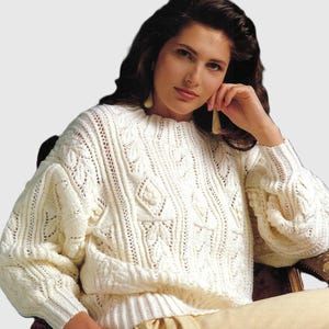 May include: Cream-colored knitted sweater with intricate cable and leaf patterns. The sweater has a round neckline and long sleeves with a textured design. The model is wearing the sweater while seated in a chair.