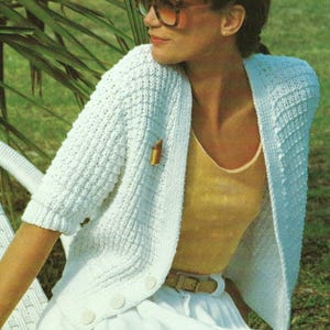 May include: A white knitted cardigan with short sleeves and a button closure. The cardigan is open in the front and has a textured stitch pattern. The model is wearing a yellow tank top and a white skirt.