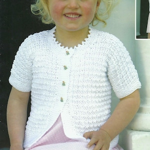 Puede incluir: Un cárdigan blanco de crochet de manga corta, con cuello festoneado y tres botones decorativos. El cárdigan se lleva sobre un vestido rosa. Los botones tienen un pequeño diseño floral.