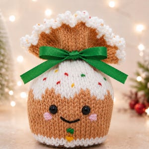 May include: A knitted pouch shaped like a gingerbread person with a white icing top, colorful sprinkles, and a green ribbon bow. The pouch features black plastic eyes, a stitched smile, pink cheeks, and a small orange detail at the bottom.