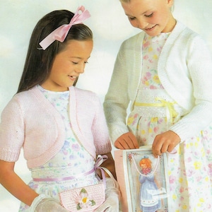 May include: Two girls wearing pink and white knitted boleros over floral dresses. The girl on the left is holding a pink crocheted purse. The girl on the right is holding a pink gift bag with a picture of a girl on it.