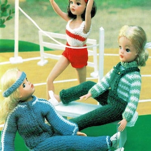 May include: Three dolls wearing knitted outfits. One doll is wearing a red and white striped tank top and red shorts. Another doll is wearing a green and white striped sweater and green pants. The third doll is wearing a blue sweater and blue pants.