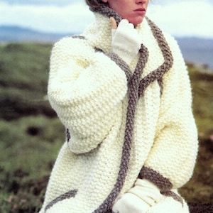 May include: A white knitted cardigan with a brown trim. The cardigan has a shawl collar and a loose fit. The model is wearing the cardigan with blue jeans.
