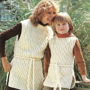 May include: Two people wearing matching cream-colored knitted vests with a cable-knit design and a matching belt. The vests are paired with brown long-sleeved shirts. The background features green foliage.