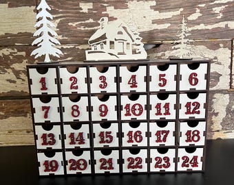 May include: A wooden advent calendar with 24 numbered drawers, each with a small handle. The numbers are in a burgundy colour. The calendar is decorated with wooden cutouts of a house and trees. The background is a rustic wooden wall.