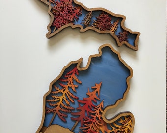 May include: Two wooden wall art pieces in the shape of the state of Michigan. The art features layered designs of trees in shades of red, orange, and yellow against a blue background. The lower piece includes bear silhouettes.