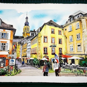 May include: Watercolor painting of a European city street scene. Buildings in shades of yellow, orange, and gray line the street. A tall church steeple rises in the background. People are walking on the street. The painting is signed "Justinian" in the lower right corner.