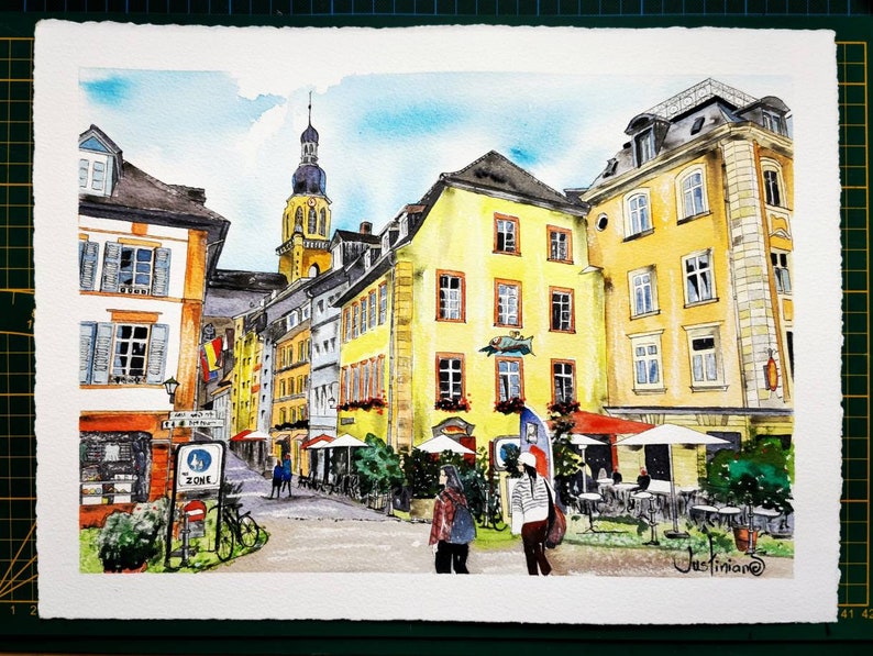 May include: Watercolor painting of a European city street scene. Buildings in shades of yellow, orange, and gray line the street. A tall church steeple rises in the background. People are walking on the street. The painting is signed "Justinian" in the lower right corner.