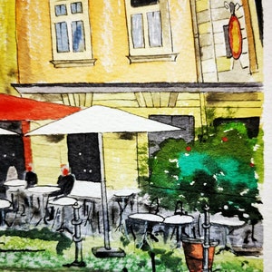 May include: Watercolor painting of a European cafe scene. The artwork depicts a yellow building with windows, a red awning, and white umbrellas over outdoor tables. Green foliage and a sign add detail. The artist's signature, "Justiniano", is in the foreground.