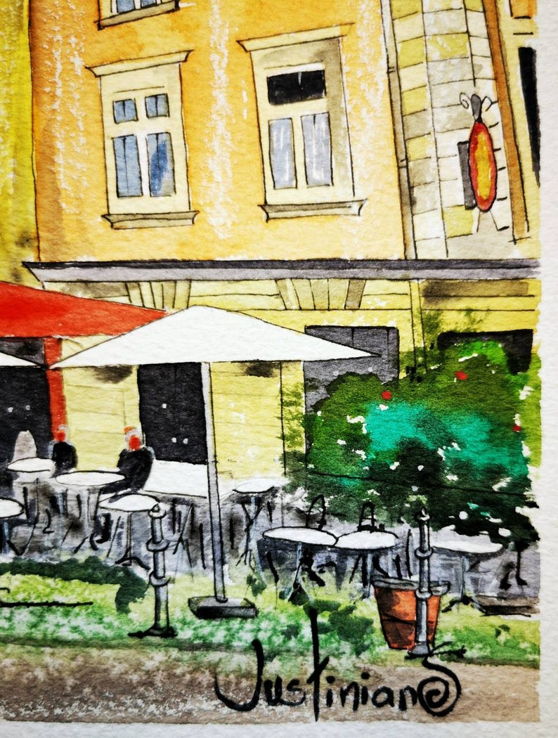 May include: Watercolor painting of a European cafe scene. The artwork depicts a yellow building with windows, a red awning, and white umbrellas over outdoor tables. Green foliage and a sign add detail. The artist's signature, "Justiniano", is in the foreground.