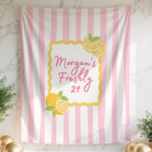 May include: A pink and white striped backdrop with the text "Morgan's Freshly 21" in pink script, framed by a yellow border and lemon illustrations. Gold and white balloons and potted plants flank the backdrop.