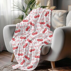 Puede incluir: Una manta blanca con un patrón de cerezas rosas y rojas. La manta está doblada y drapeada sobre un sillón blanco.