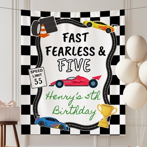 May include: A rectangular backdrop featuring a black and white checkered border, a central white area with a winding road, and the text FAST FEARLESS & FIVE. It includes illustrations of race cars, a trophy, tires, and a SPEED LIMIT 55 sign.