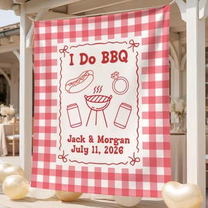 May include: A rectangular fabric banner with a red and white gingham border. The center features the red text I Do BBQ, a hot dog, a diamond ring, a grill, and two cans. Below are the names Jack & Morgan and the date July 11, 2026.