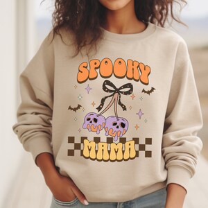 Könnte beinhalten: Beiges Sweatshirt mit den Worten "SPOOKY MAMA" in orangefarbener und gelber Retro-Schrift. Die Grafik zeigt lila Totenkopf-Kirschen mit einer schwarzen Schleife, Fledermäusen und Sternen. Unten befindet sich ein Schachbrettmuster.