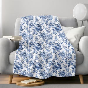 Op de afbeelding: Een witte deken met een blauw bloemenpatroon. De deken heeft een herhalend patroon van blauwe rozen en bladeren op een witte achtergrond.