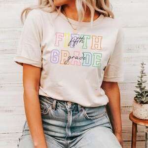 K&ouml;nnte beinhalten: Ein wei&szlig;es T-Shirt mit dem Text "FIFTH grade" in Regenbogenfarben. Der Text ist in einer fetten, blockartigen Schrift.