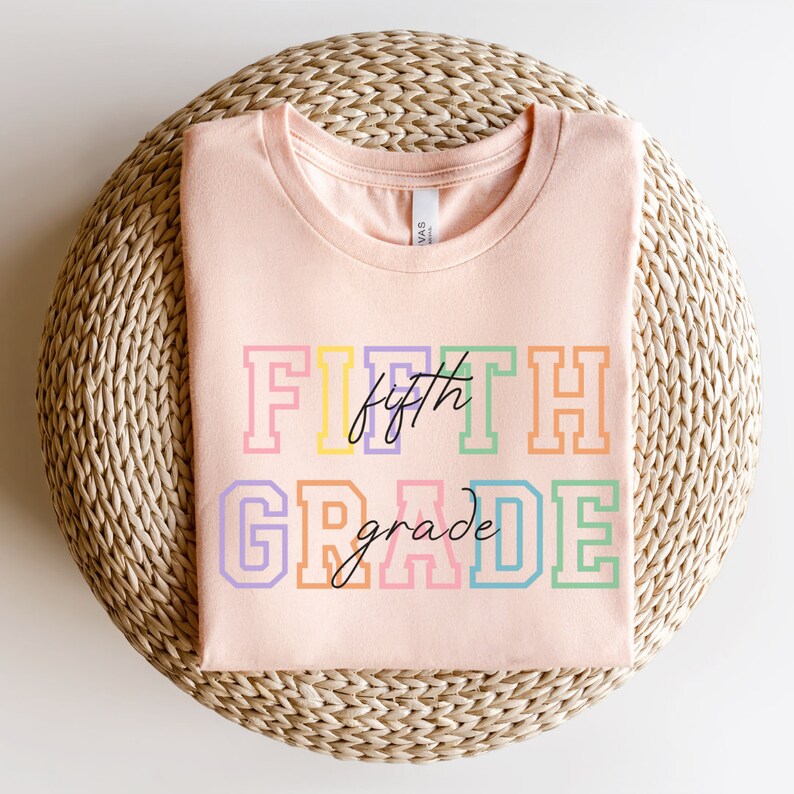 K&ouml;nnte beinhalten: Ein rosa T-Shirt mit dem Text "FIFTH grade" in Regenbogenfarben. Der Text ist schwarz umrandet und das Wort "fifth" ist kursiv geschrieben.