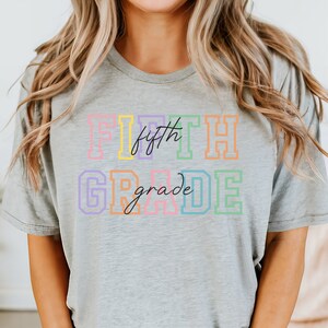 K&ouml;nnte beinhalten: Ein graues T-Shirt mit dem Text "FIFTH grade" in Regenbogenfarben. Das Wort "fifth" ist kursiv geschrieben.