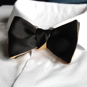 Puede incluir: Una pajarita negra con ribete dorado, colocada sobre el cuello de una camisa blanca. La pajarita está hecha de un material brillante y está cuidadosamente atada. El ribete dorado añade un toque de elegancia.