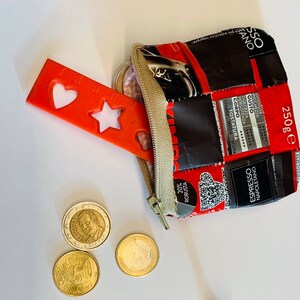 Può includere: Una piccola borsa rossa con chiusura a zip, realizzata con imballaggi di caffè riciclati. La borsa è aperta e contiene una forma di cuore e stella in plastica rossa, due monete da euro e una moneta dorata. La borsa è su una superficie bianca.