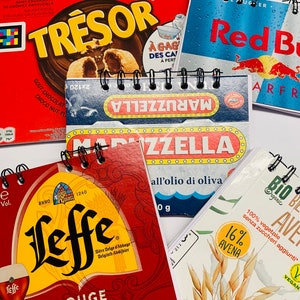 Pode incluir: Uma coleção de embalagens de produtos coloridos que apresentam várias marcas. As embalagens incluem uma caixa vermelha com a palavra "TRESOR" em letras amarelas, uma caixa azul e branca com a palavra "MARUZZELLA" em letras brancas, uma caixa vermelha e amarela com a palavra "Leffe" em letras pretas, uma caixa vermelha e azul com as palavras "Red Bull" em letras brancas, e uma caixa branca com as palavras "Bio Ave" em letras pretas.