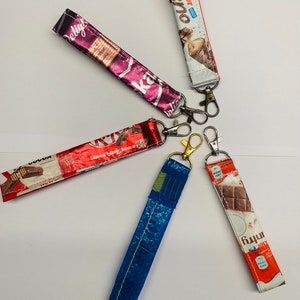 May include: Five colorful keychains made from recycled candy bar wrappers. The keychains feature different candy bar brands, including Kinder Bueno, Twix, and a red wrapper with the text "Have a break, have a Kit Kat".