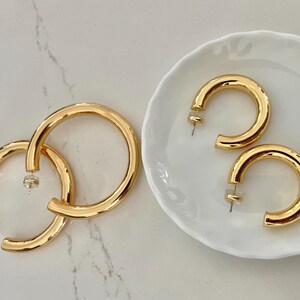 Puede incluir: Dos pares de pendientes de aro dorados. Un par es más grande y descansa sobre una superficie junto a un par de pendientes más pequeños exhibidos en un plato blanco festoneado. Los pendientes tienen un acabado brillante y pulido.