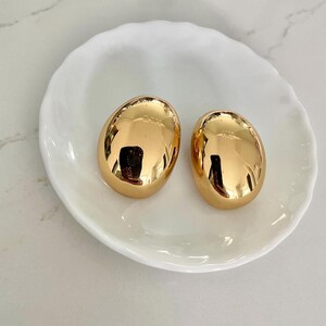 Puede incluir: Dos pendientes dorados de forma ovalada con un acabado brillante. Los pendientes se muestran sobre un plato de cerámica blanco de forma orgánica. Los pendientes reflejan el entorno.