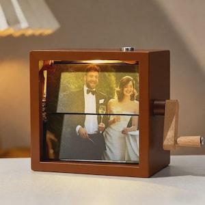 May include: A brown wooden photo cube with a hand crank, displaying a wedding photo. The cube has a warm light inside, illuminating the couple. The crank is made of wood and has a small wooden handle.